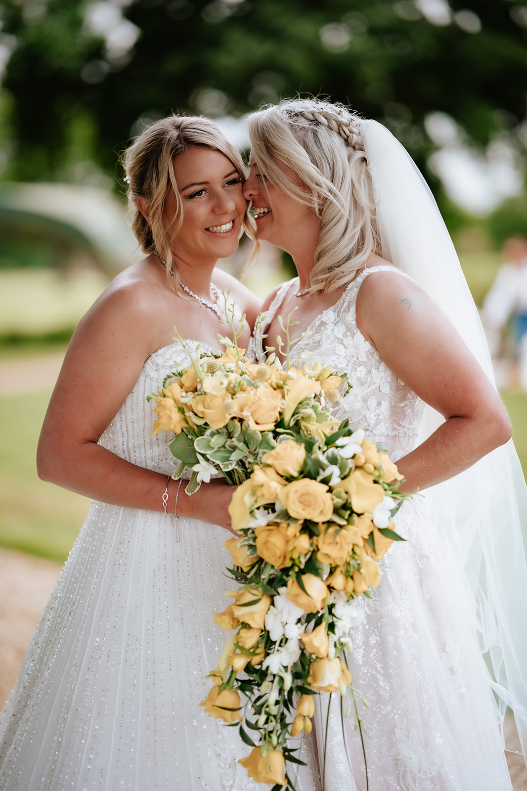 Two brides embracing with yellow rose bouquet in the parkland grounds at Gosfield Hall wedding — LGBTQ+ wedding photography by Lily & White