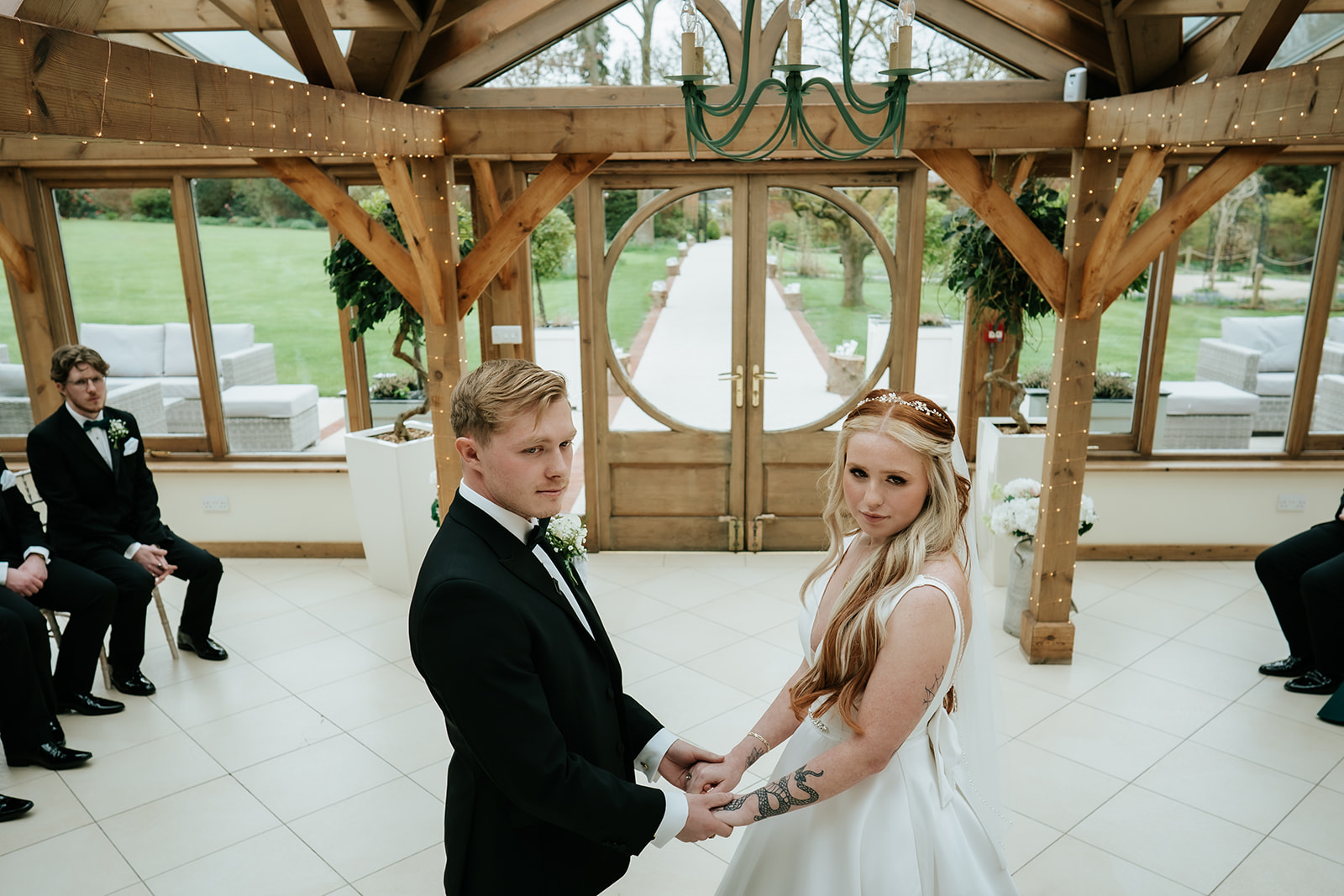 Olivia and Alfie holding hands at the altar during their wedding ceremony in the Orangery at Gaynes Park, the circular window and Long Walk visible through the glass doors behind them – wedding photography by Tel, Lily & White