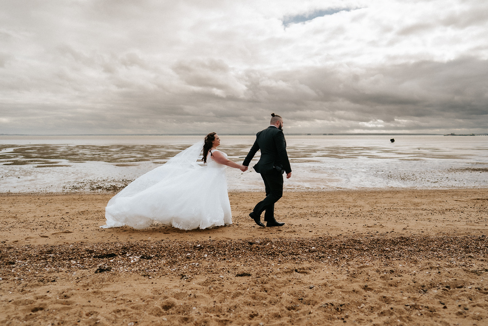 Bride and groom walking hand-in-hand along the beach at Southend-on-Sea, Essex, with dramatic estuary skies and the Thames in the background — editorial wedding photography by Lily & White