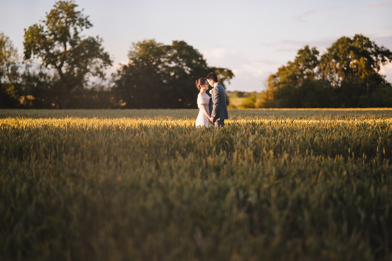 Yiran and Jeffrey holding hands in the wheat field behind the Essex Barn at Blake Hall during golden hour, warm evening sun flooding across the Essex countryside – wedding photography by Tel, Lily & White