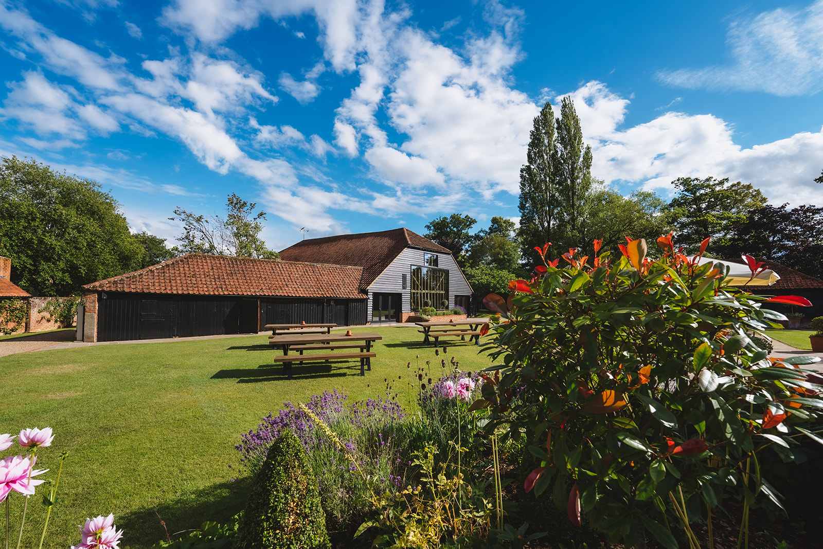 The grounds of Blake Hall wedding venue in Essex, with the Hay Barn visible beyond colourful cottage gardens and picnic benches on the lawn