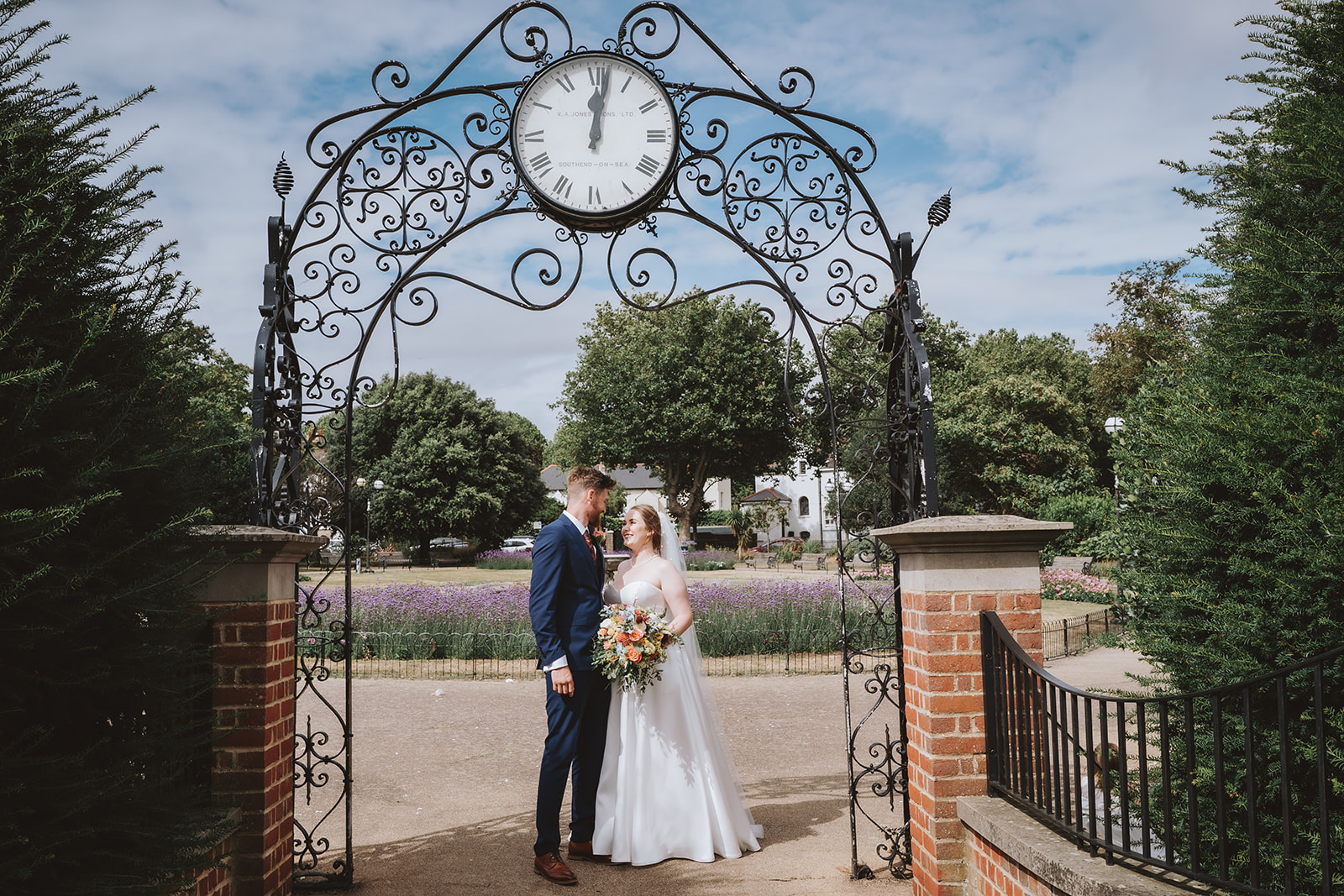 Bride and groom sharing an intimate moment beneath the iconic ornate clock arch in Southend, with lavender gardens behind them — editorial wedding portrait by Lily & White Essex wedding photographer