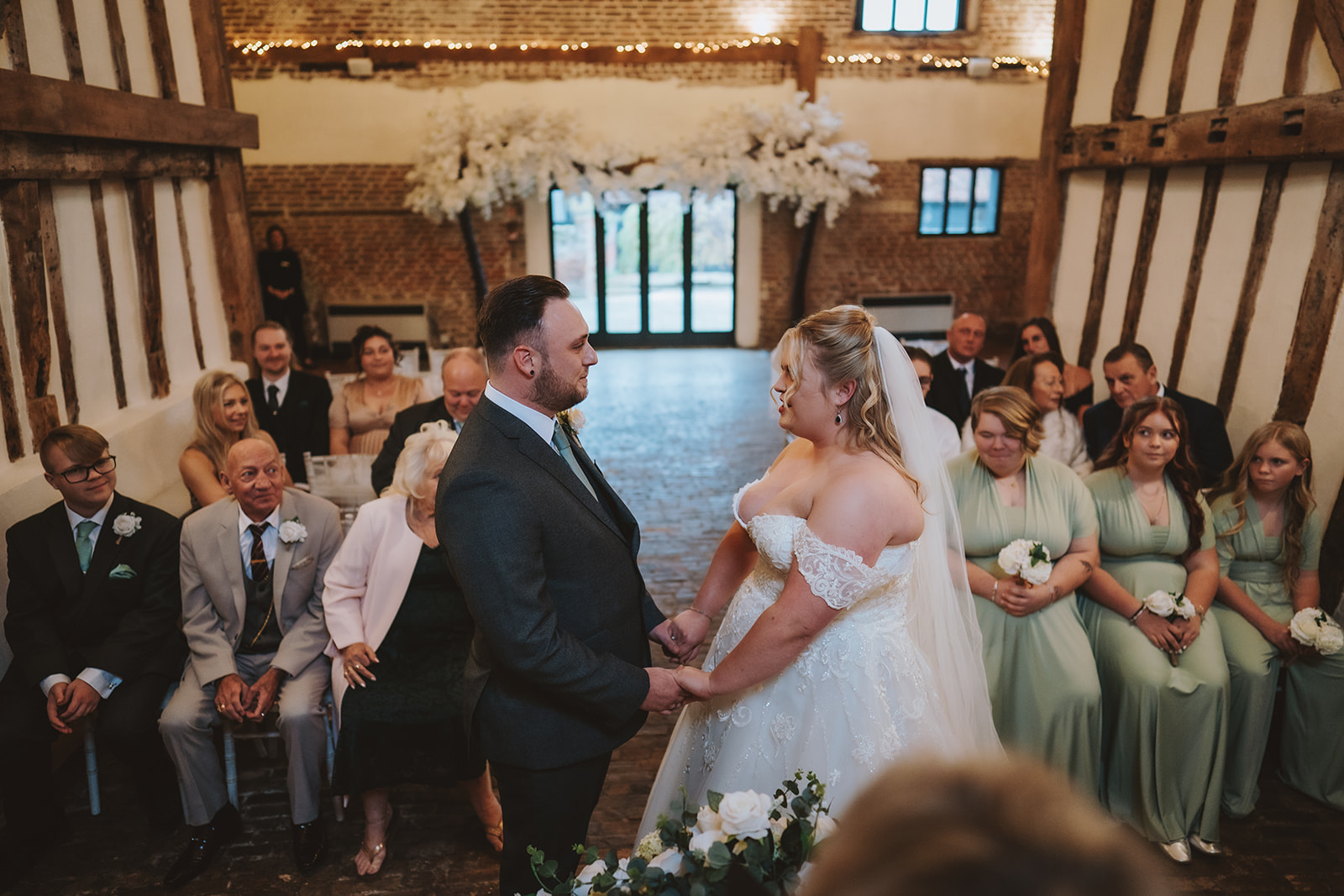 A bride and groom exchange vows inside the Coach House Barn at Leez Priory wedding venue in Little Leighs, Essex. The couple hold hands at the altar beneath exposed timber beams and fairy lights, surrounded by guests and sage green bridesmaids. Photographed by Tel, Lily & White Photography, Leez Priory wedding photographer.