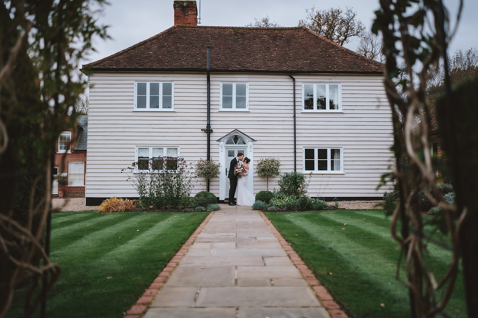 Abbey and Charlie kissing at the entrance to the bridal cottage at Blake Hall, manicured lawn and pathway leading up to the white weatherboarded building – wedding photography by Tel, Lily & White