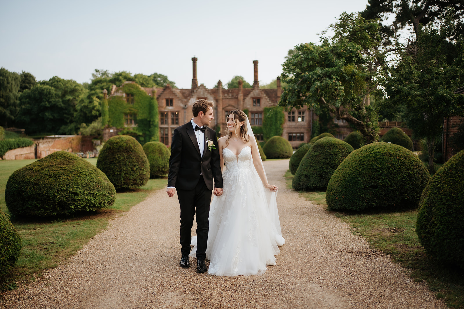 A bride in a lace strapless gown and a groom in a black tuxedo holding hands and walking along a gravel path toward a stately manor.