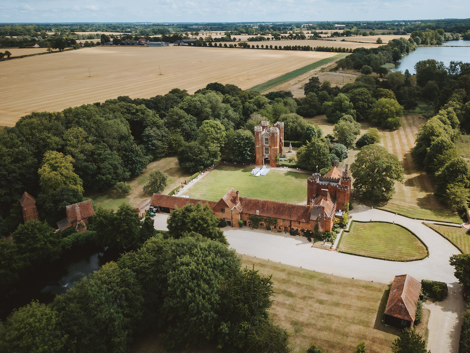 Aerial drone photograph of Leez Priory wedding venue in Little Leighs, Essex, showing the full Tudor estate including the Great Tower, the Coach House, surrounding parkland and lakes, with open Essex countryside and farmland stretching to the horizon. Photographed by Tel, Lily & White Photography, Leez Priory wedding photographer.