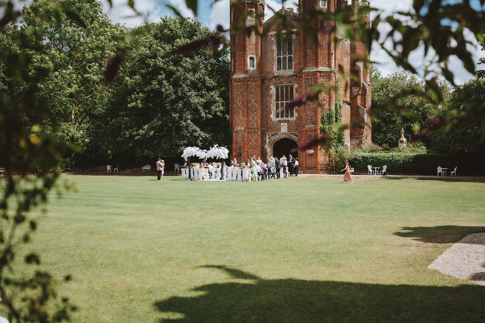 A wide view of an outdoor wedding ceremony taking place on the lawn in front of the Great Tower at Leez Priory, Little Leighs, Essex. Guests are seated in rows facing the Tudor brick tower on a sunny summer day. Photographed by Tel, Lily & White Photography, Leez Priory wedding photographer.