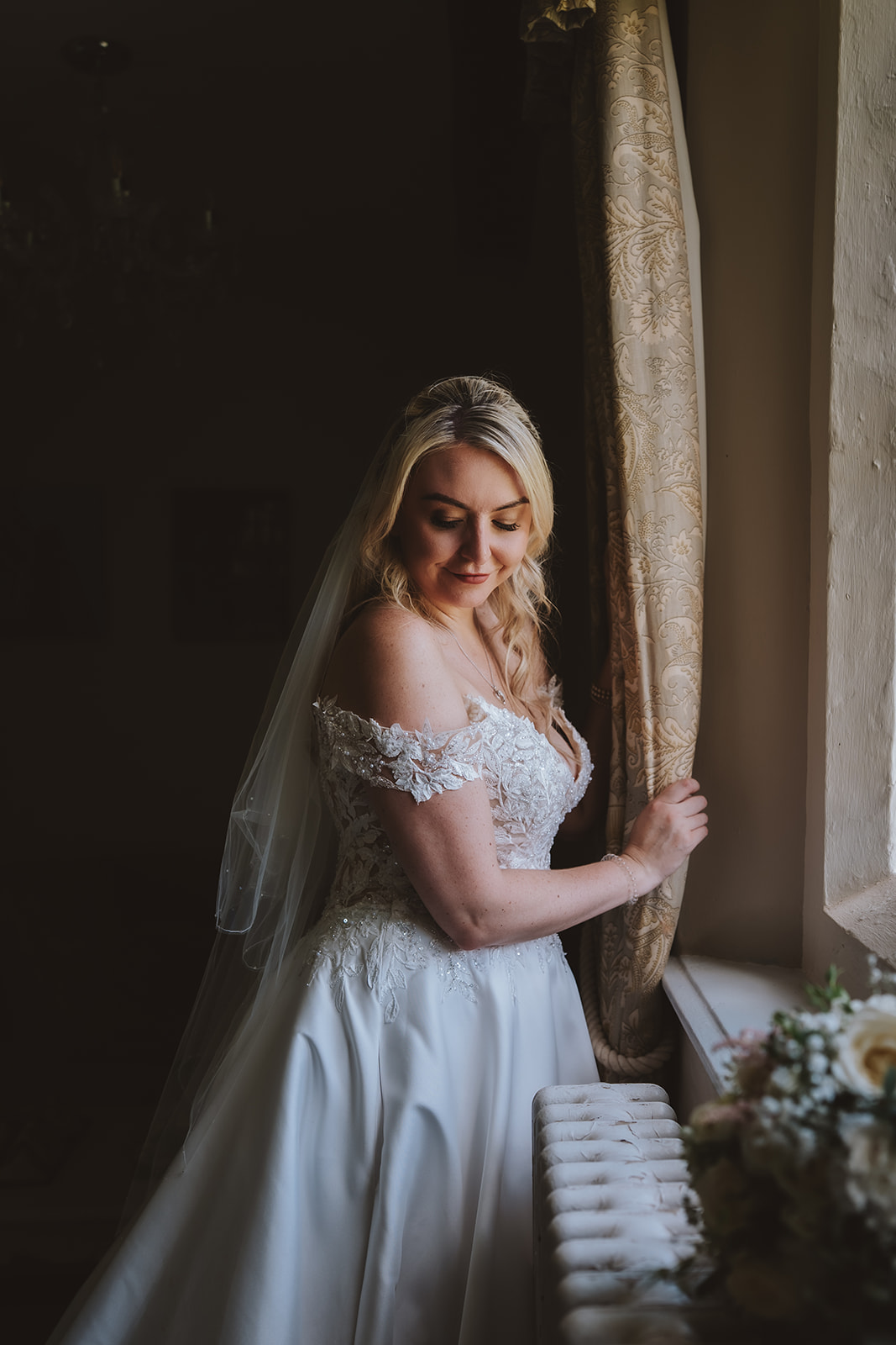 A bride stands in soft window light inside Leez Priory, Little Leighs, Essex, her off-shoulder lace wedding dress and veil catching the warm glow from the old stone window frame. Photographed by Tel, Lily & White Photography, Leez Priory wedding photographer.