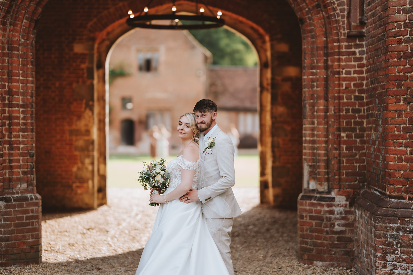 A bride and groom stand together framed within the ancient Tudor brick archway of the carriageway at Leez Priory, Little Leighs, Essex. The depth of the arch and the courtyard visible beyond creates a natural frame around the couple. Photographed by Tel, Lily & White Photography, Leez Priory wedding photographer.
