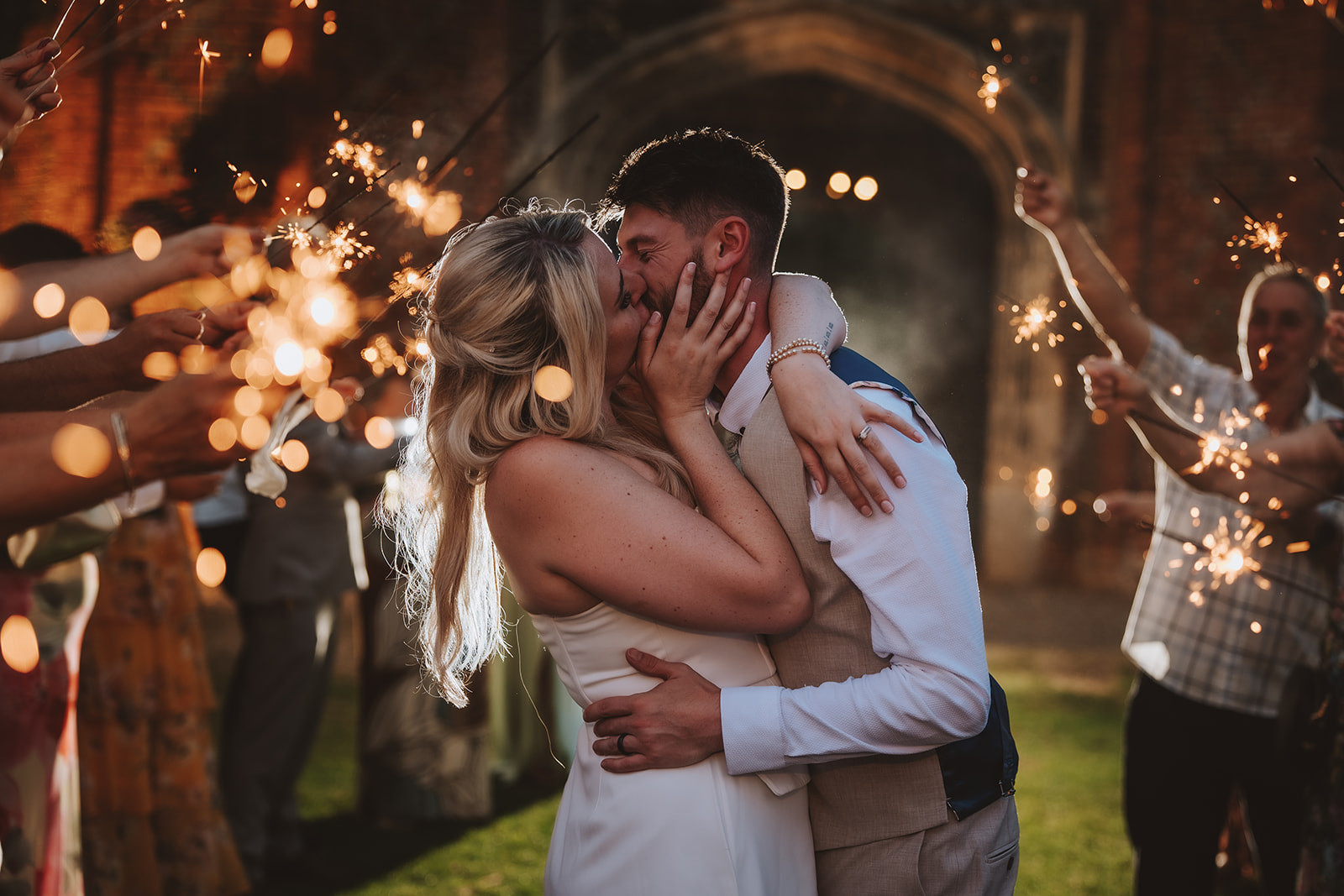 A bride and groom share a passionate kiss during a sparkler send-off beneath the Tudor carriageway arch at Leez Priory, Little Leighs, Essex. Guests surround the couple holding lit sparklers, their light reflected in bokeh circles against the ancient red brick archway. Photographed by Tel, Lily & White Photography, Leez Priory wedding photographer.