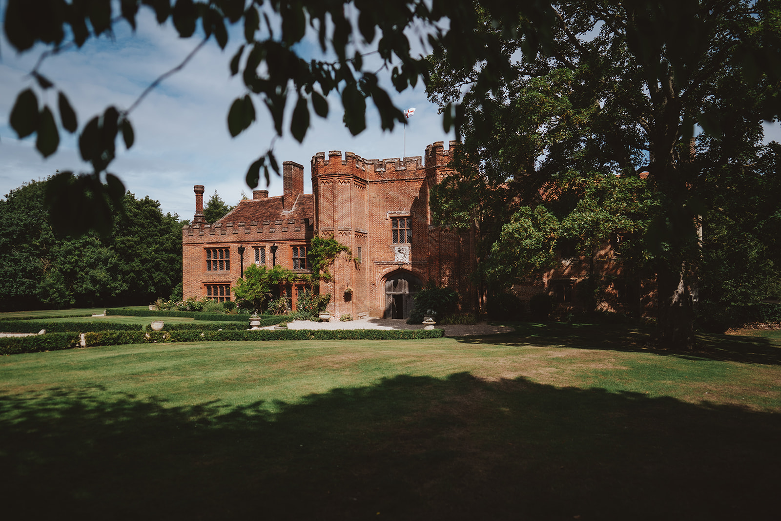 The Tudor redbrick facade and castellated towers of Leez Priory wedding venue framed by mature trees and manicured parkland in Essex — editorial wedding photography by Lily & White