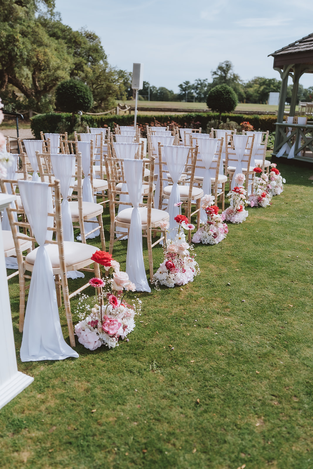 A minimalist and elegant outdoor wedding ceremony setup in Essex, featuring white chair sashes and vibrant floral arrangements on a sunny day.