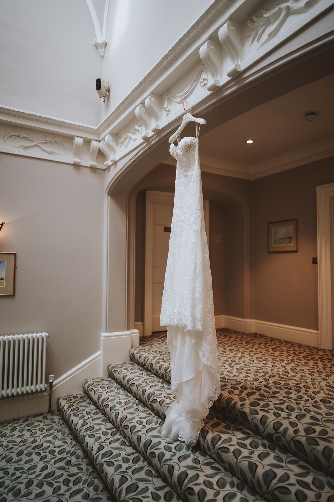 Wedding dress hanging in the elegant Georgian staircase at Wivenhoe House with period plasterwork and patterned carpet — detail photography by Lily & White