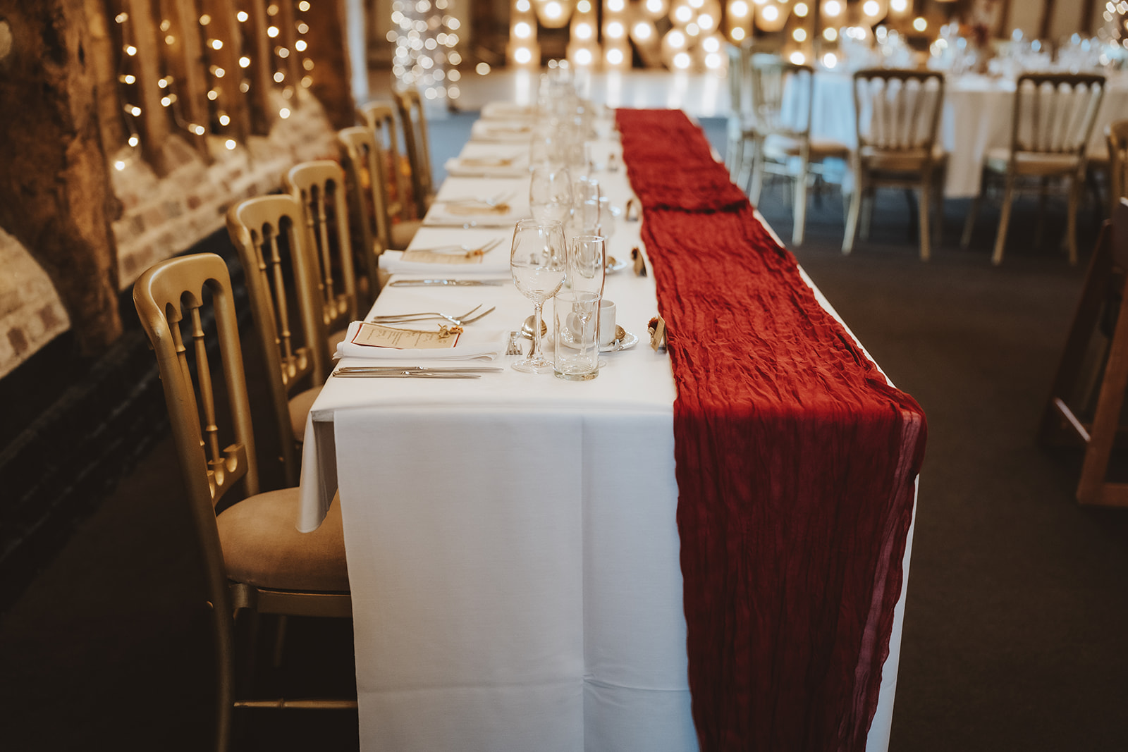 a long top table with deep red velvet table runner and gold place settings in the Essex barn
