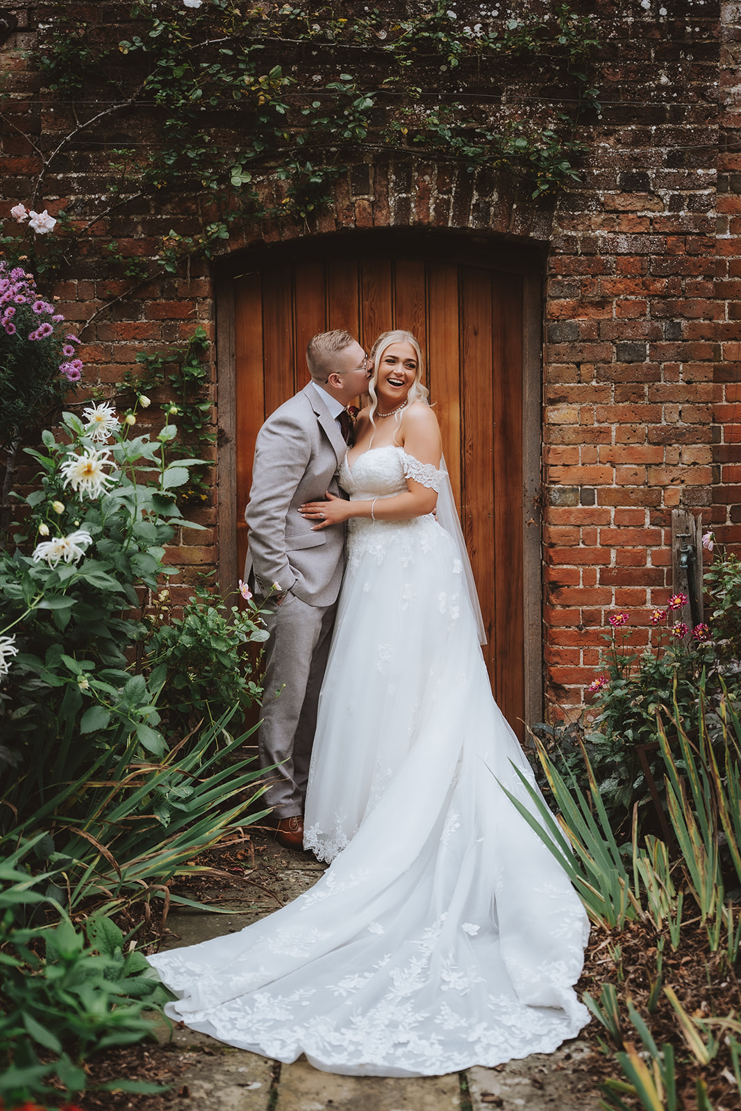 Holly and Billy laughing together in the doorway of a brick arch at Blake Hall, dahlias and climbing plants framing them.