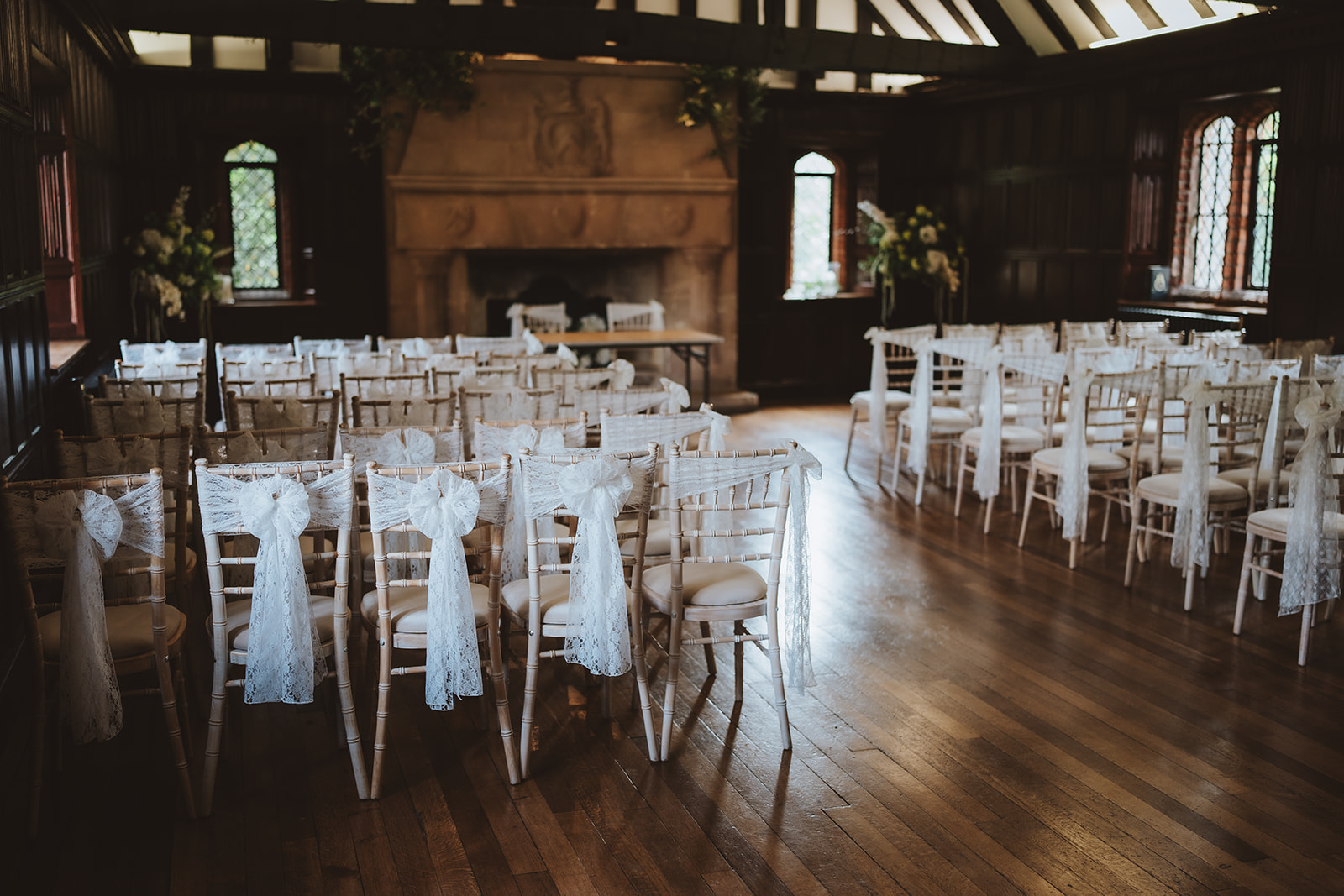 The Great Hall at Leez Priory wedding venue in Little Leighs, Essex, dressed for a wedding ceremony with rows of white chiavari chairs, white sash bows and a stone fireplace. Photographed by Tel, Lily & White Photography, Leez Priory wedding photographer.