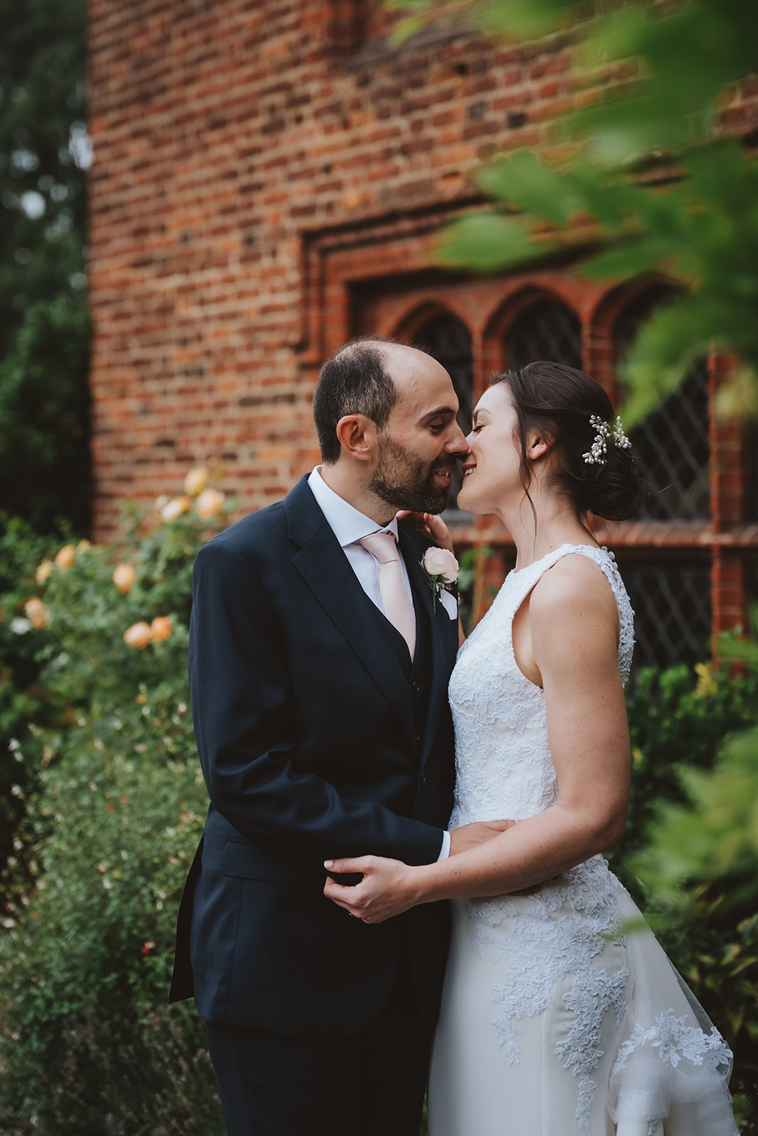 A bride and groom share an intimate moment in the gardens beside the Tudor brick walls of Leez Priory, Little Leighs, Essex. The groom whispers to the bride as she laughs, surrounded by climbing roses and lush green foliage. Photographed by Tel, Lily & White Photography, Leez Priory wedding photographer.