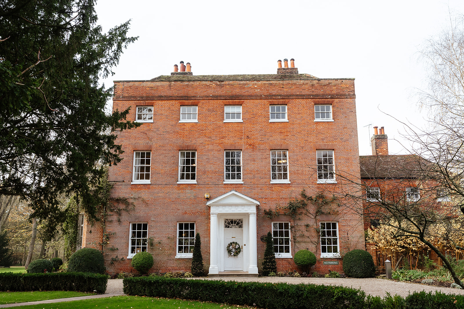 The Georgian red brick facade of Mulberry House wedding venue in Essex, decorated with a winter wreath, surrounded by mature trees in late autumn — photographed by Lily & White
